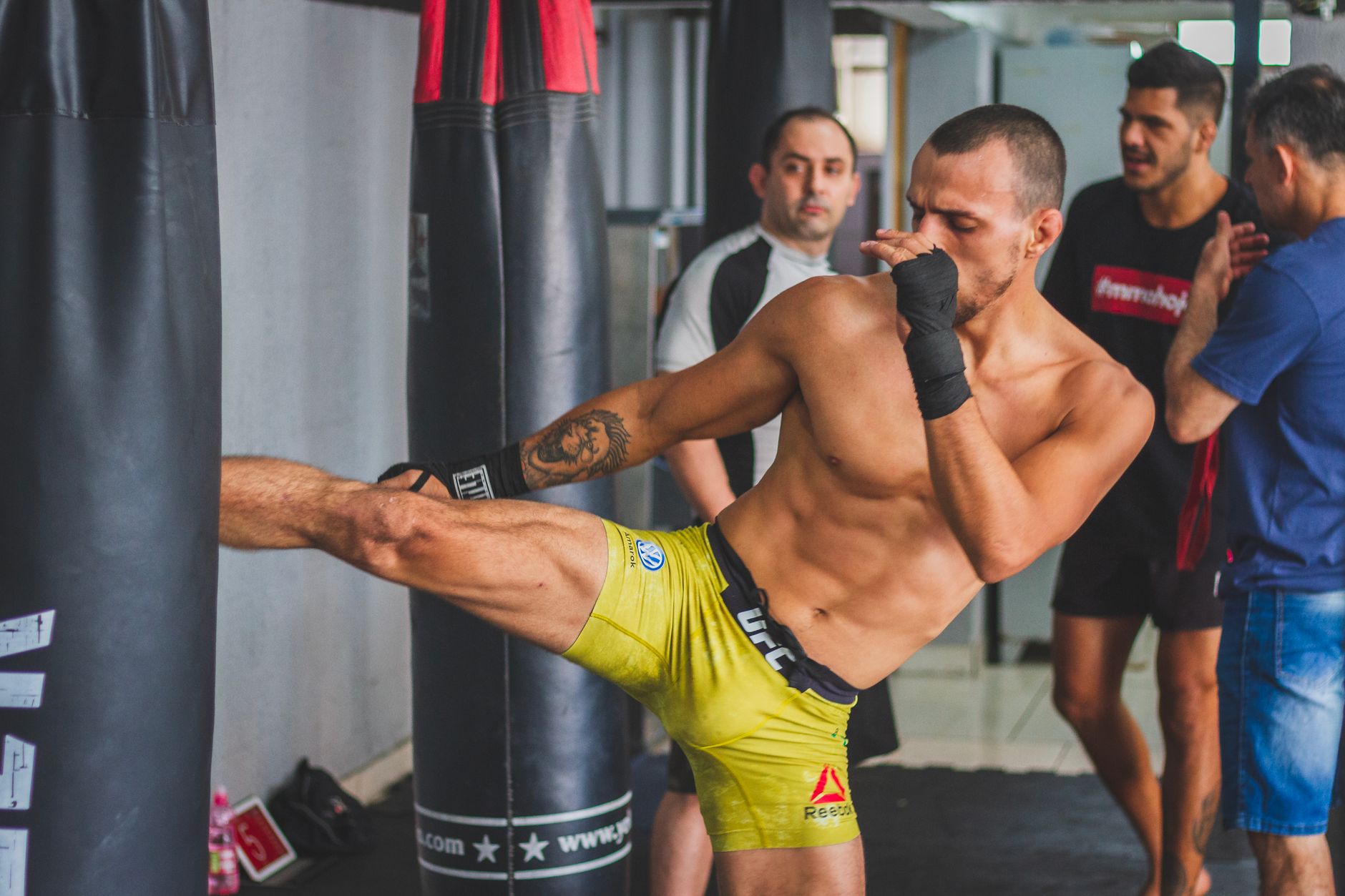 A martial artist executes a kick on a heavy bag during indoor training, surrounded by others.