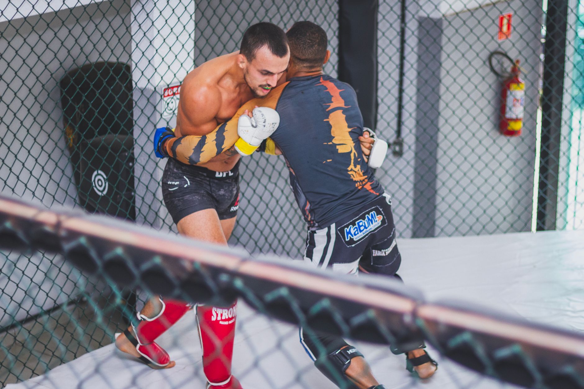 Two male athletes engaged in a competitive mixed martial arts training session in an indoor cage.
