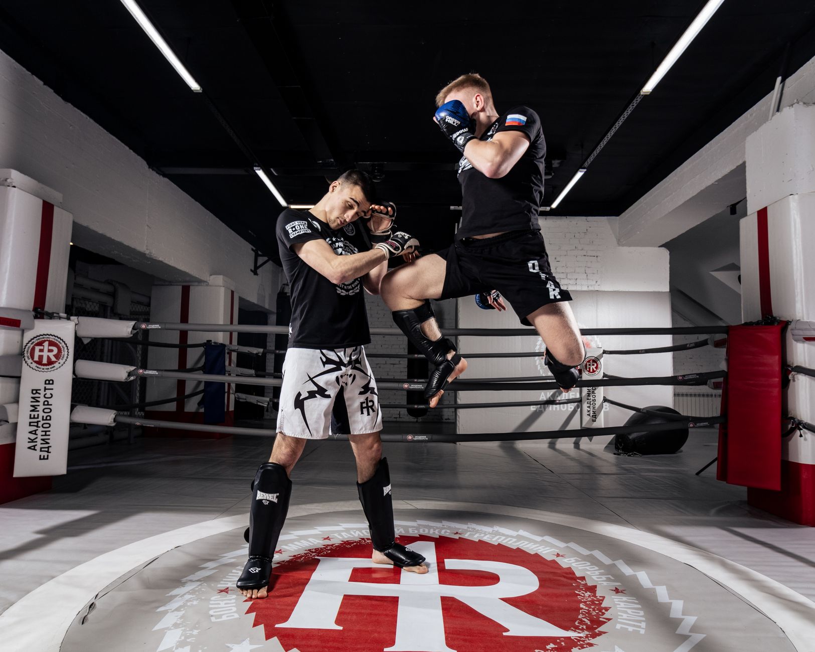 Two men practice dynamic martial arts in a well-lit gym setting.