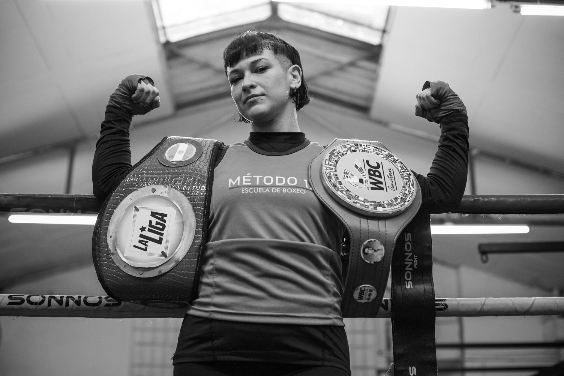 Female boxer proudly displays championship belts in an indoor gym setting.