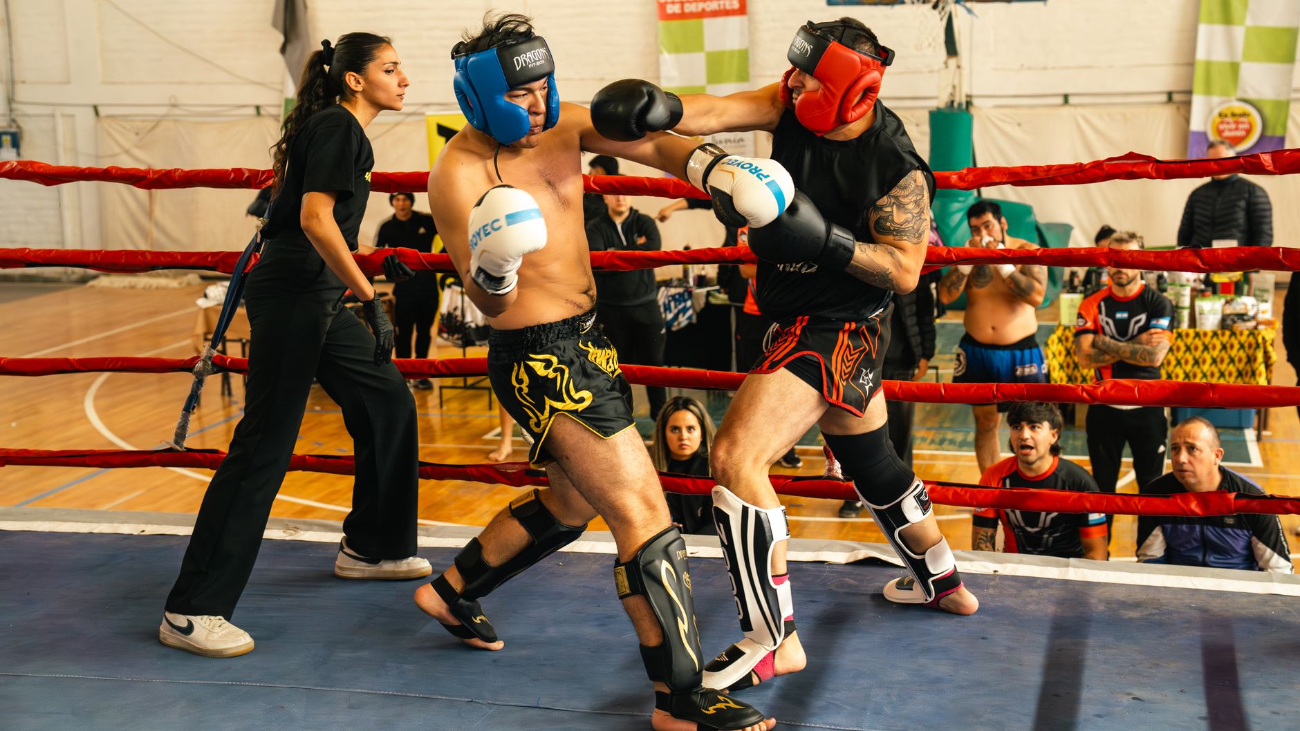 Two fighters engaged in a dynamic Muay Thai match with a referee overseeing in an indoor arena.