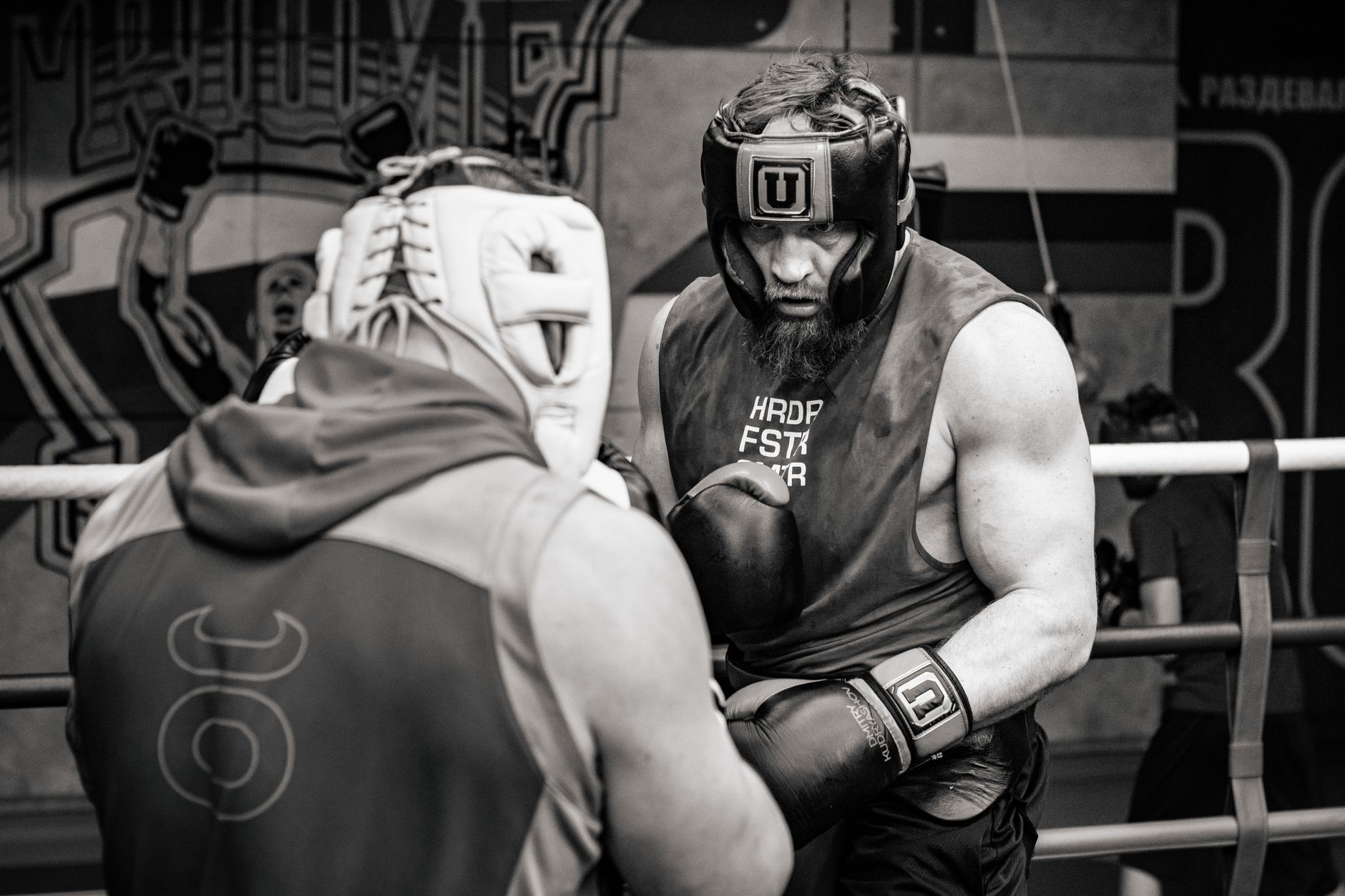 Two muscular men sparring in a boxing ring, wearing protective gear, in a gritty gym atmosphere.