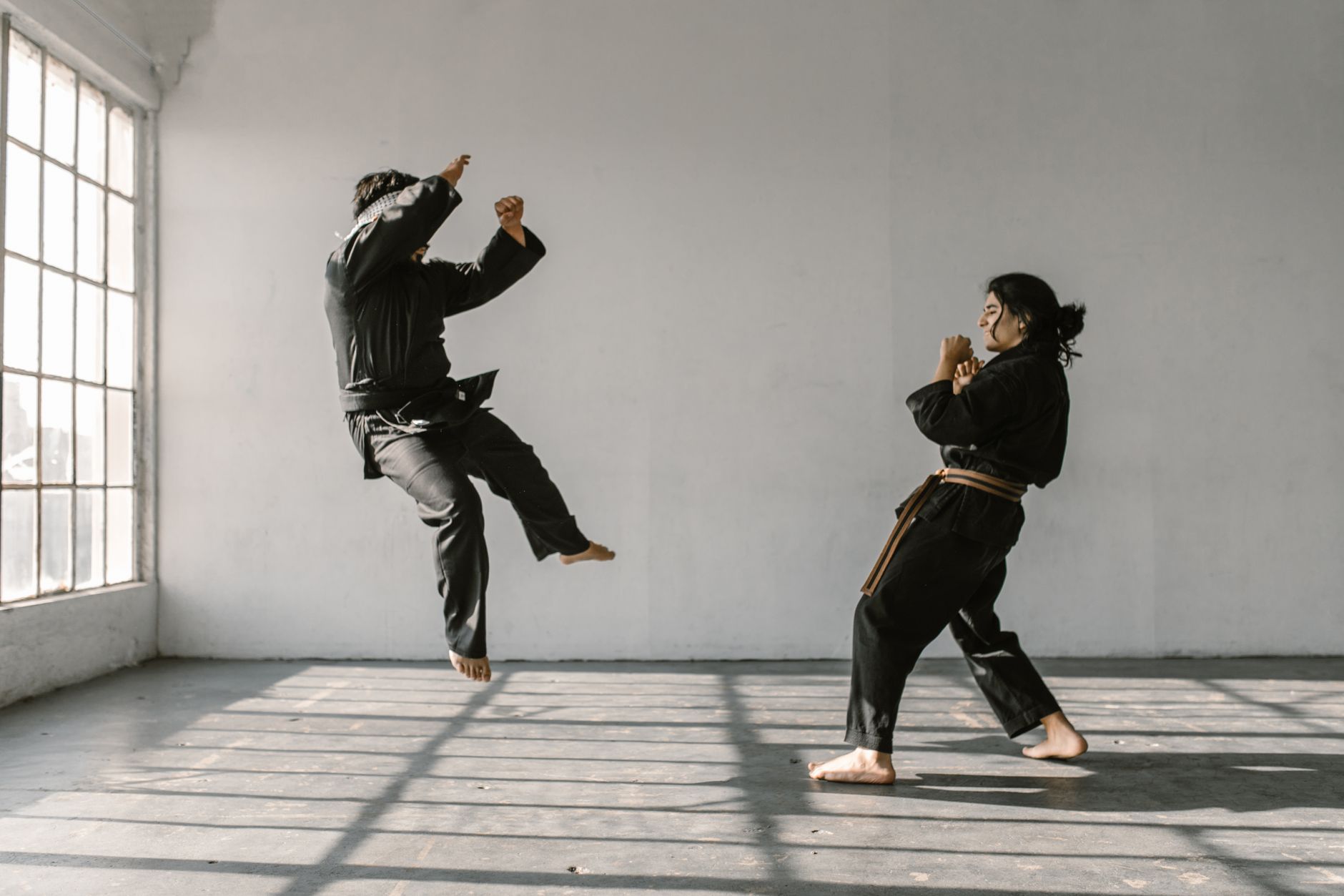 Two martial artists in black gear practice sparring in a well-lit dojo.