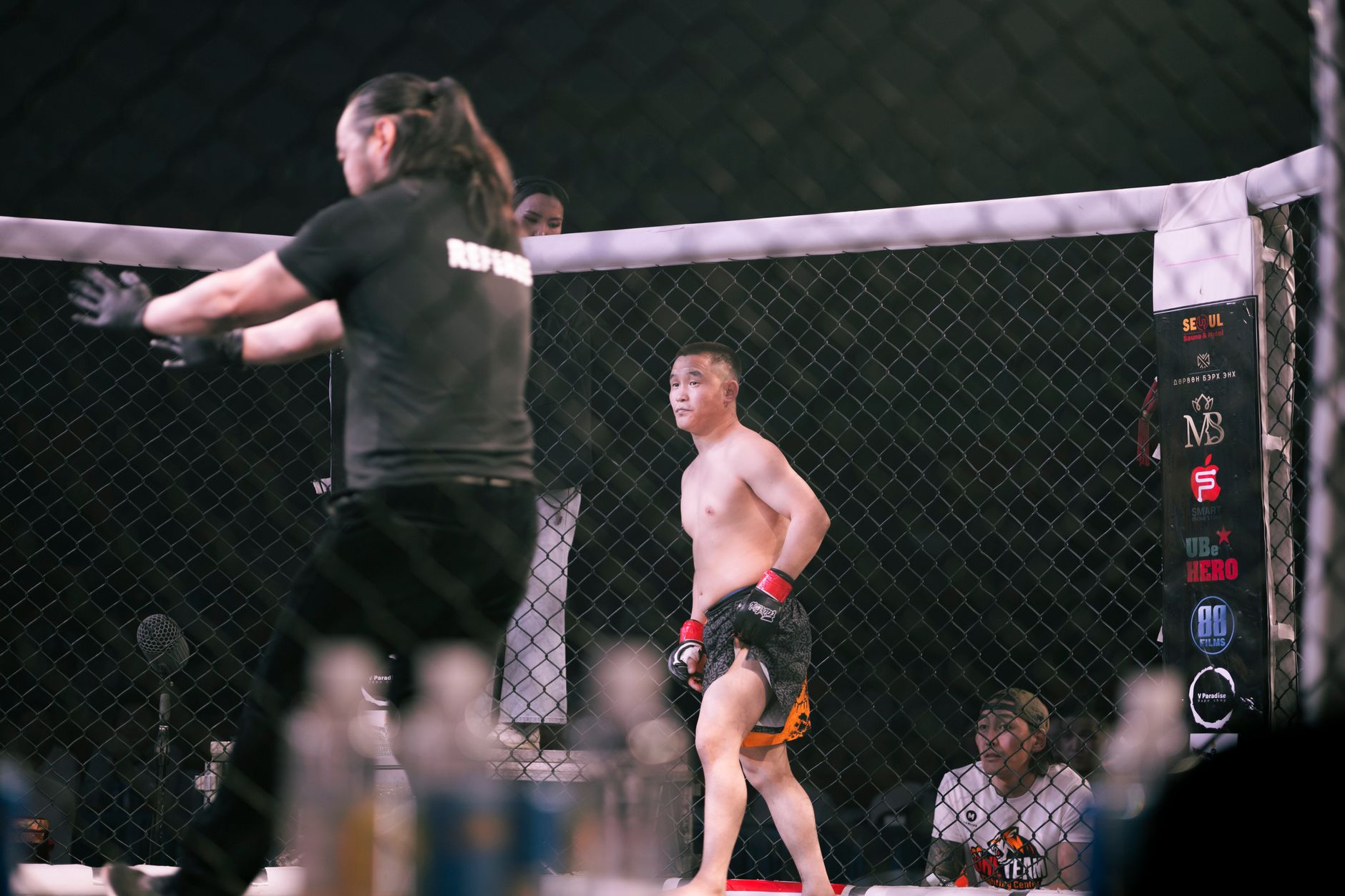 Mixed martial arts fight featuring two opponents and a referee in a caged arena.