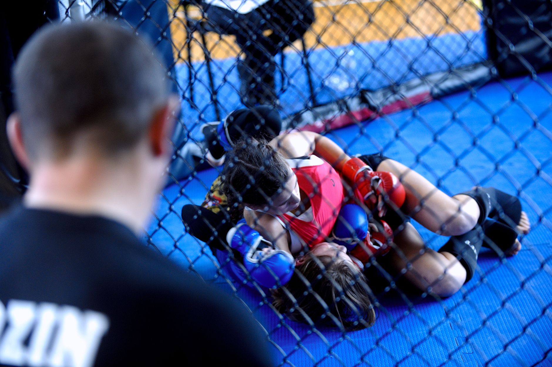 Two women competing in a mixed martial arts training session inside a gym cage with a coach observing.