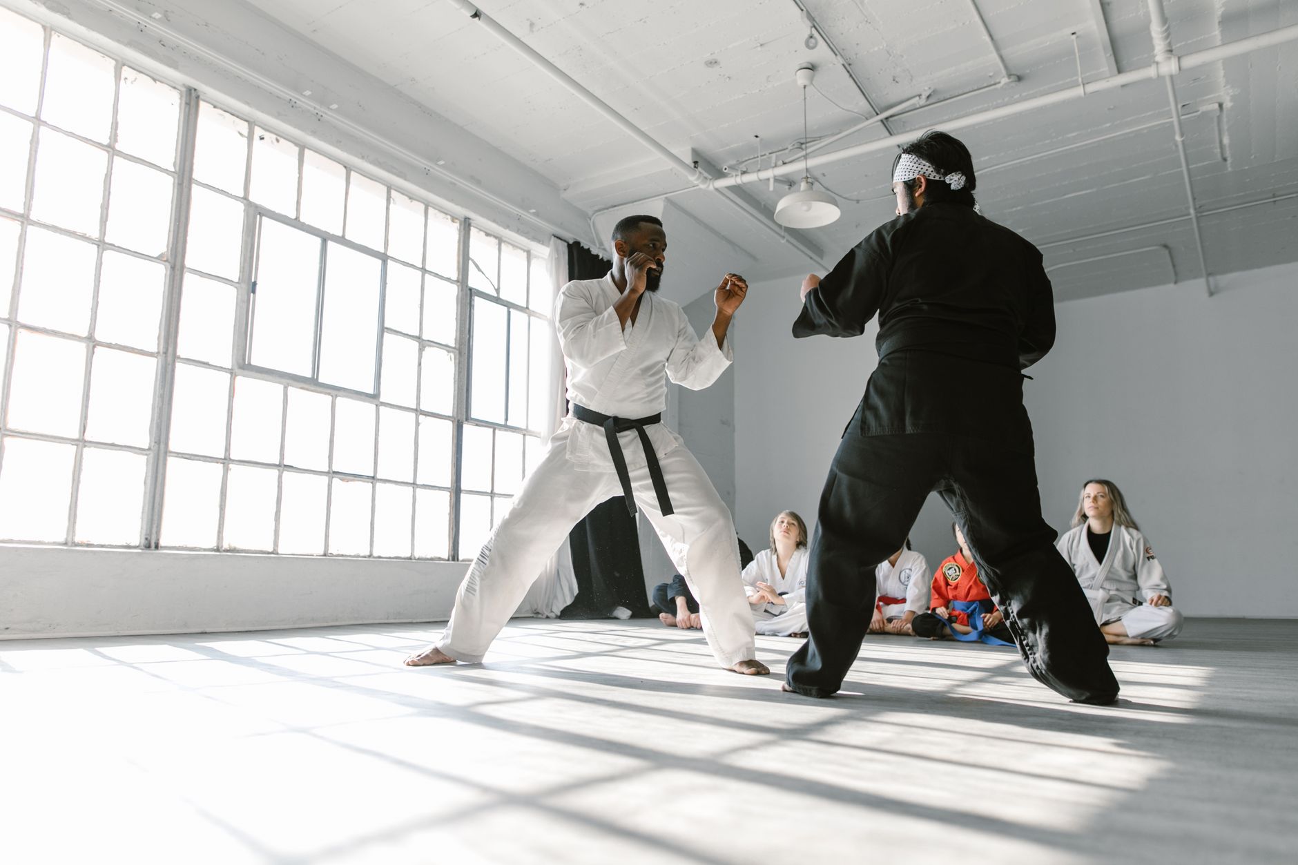 Two martial artists sparring in a dojo with a group watching attentively.