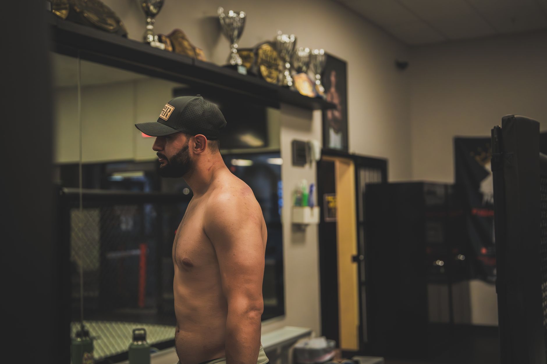 Shirtless man wearing a cap in a gym's locker room, next to trophies.