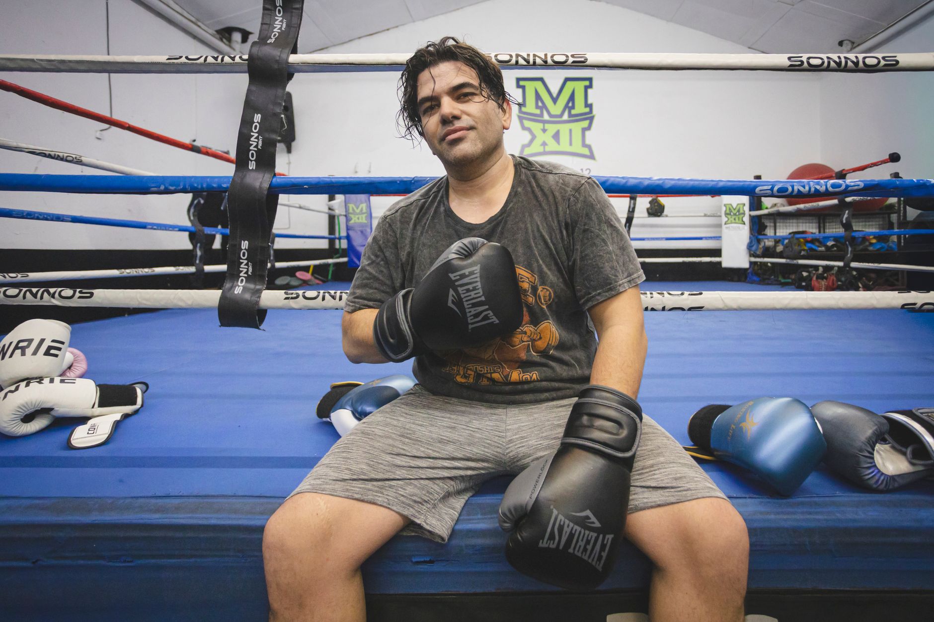 A male boxer sitting in a boxing ring after training, wearing gloves and a sweaty shirt.