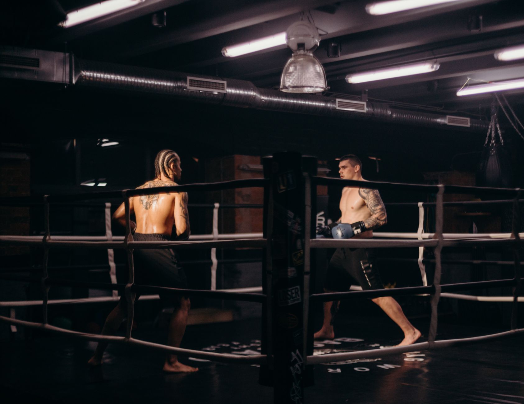 Two boxers engage in an intense match inside a dimly lit gym boxing ring.