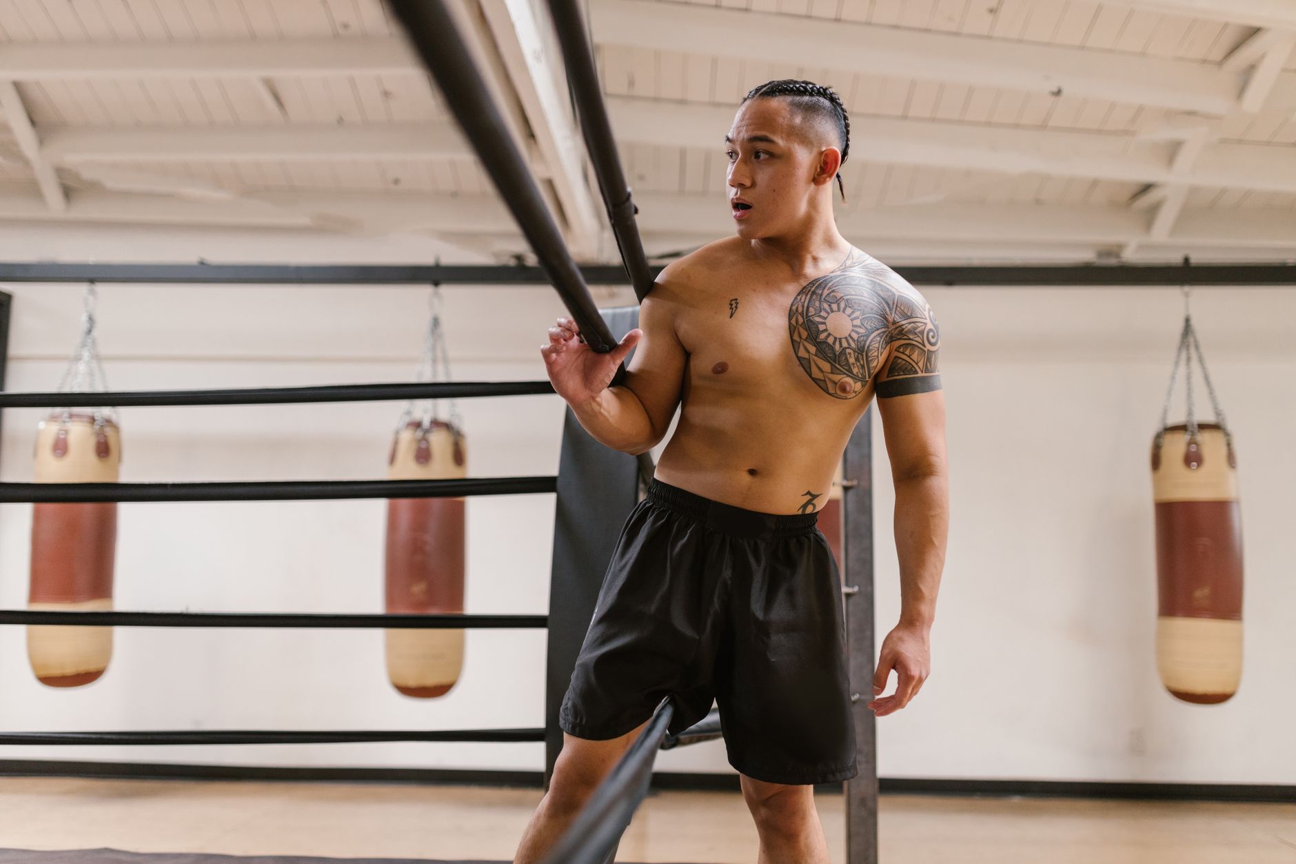 Shirtless martial artist training in a gym with punching bags and a focused expression.