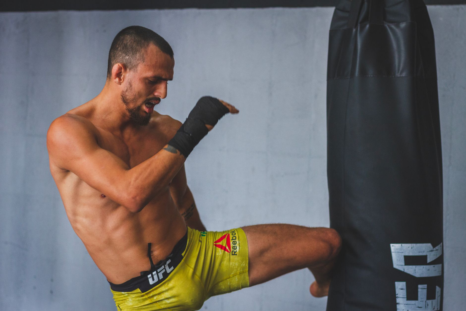 Muscular man practicing martial arts, shirtless, kicking a punching bag indoors.