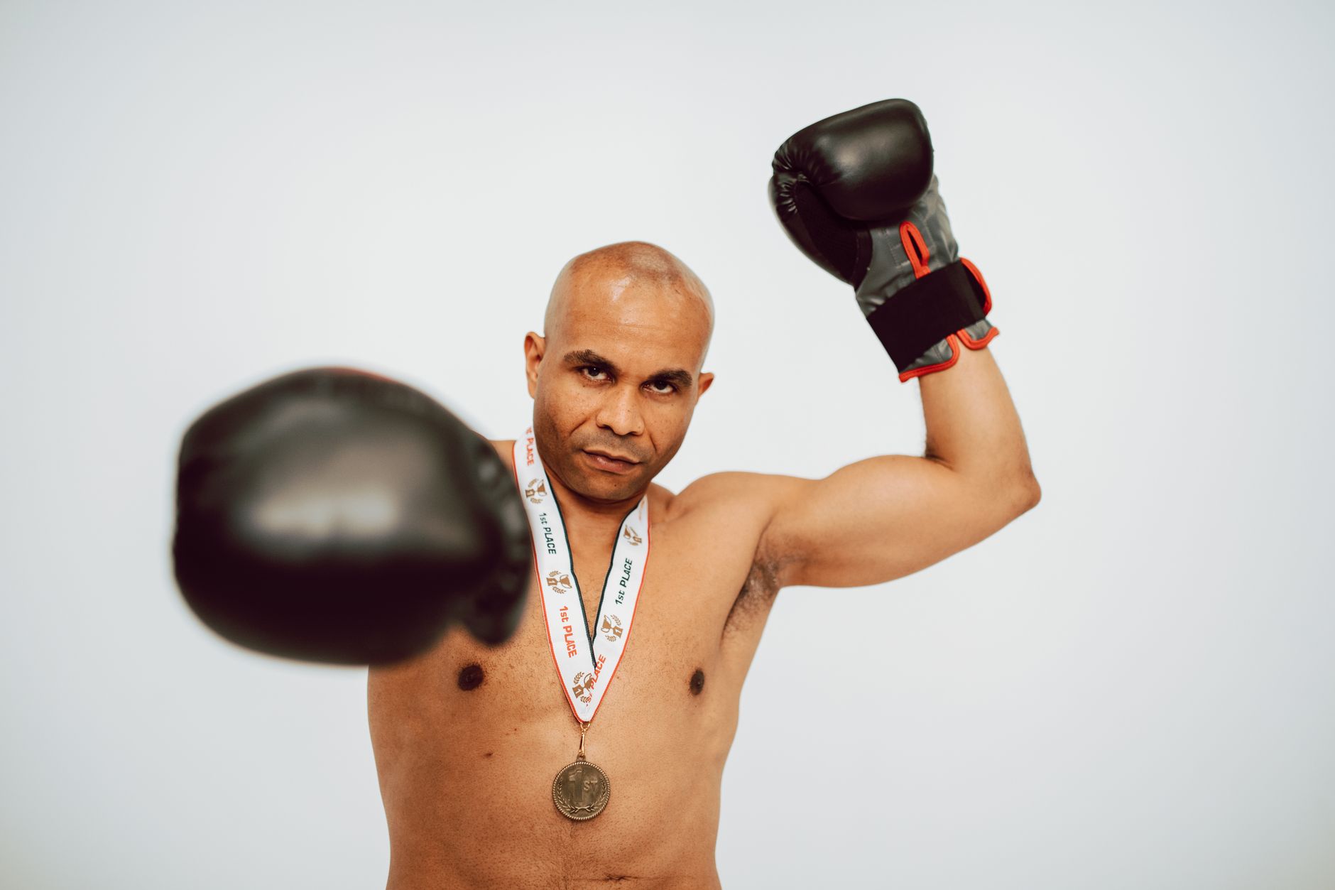 Shirtless bald man wearing boxing gloves and medal, photographed in action pose.