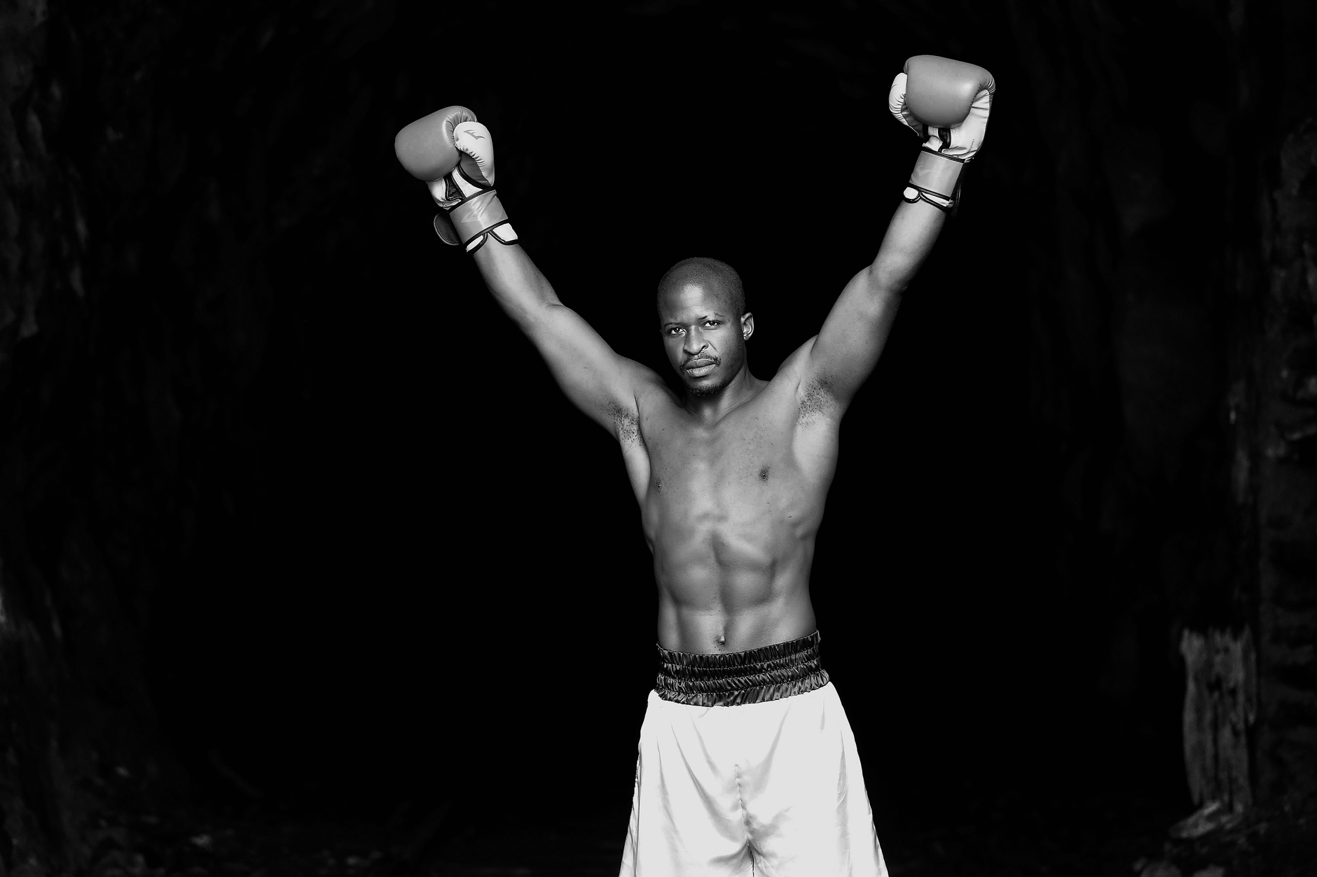 A strong, shirtless boxer triumphantly raising his gloved hands in a dramatic grayscale photograph.