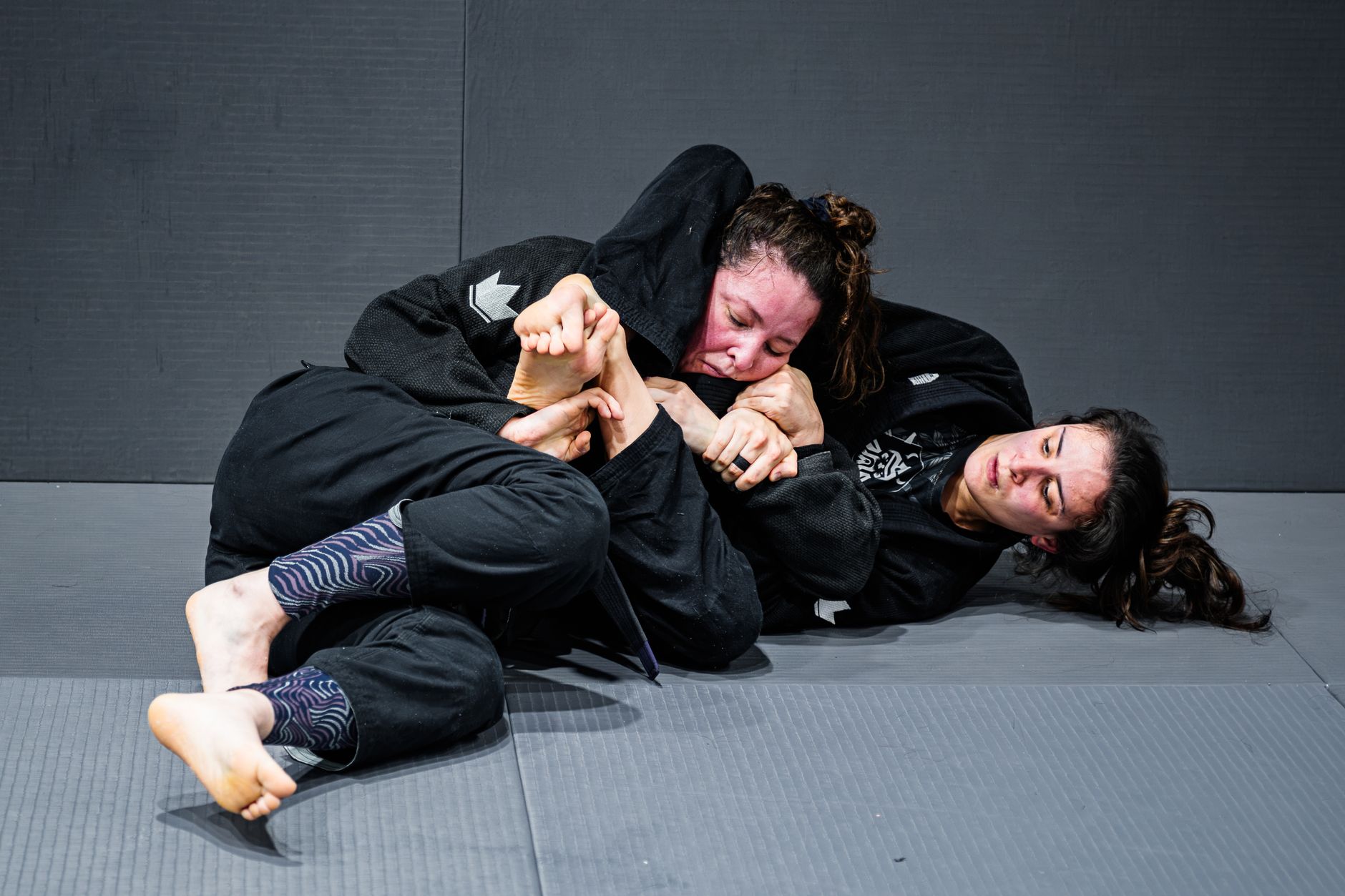 Two female martial artists engaged in a competitive Brazilian Jiu-Jitsu match on the mat indoors.